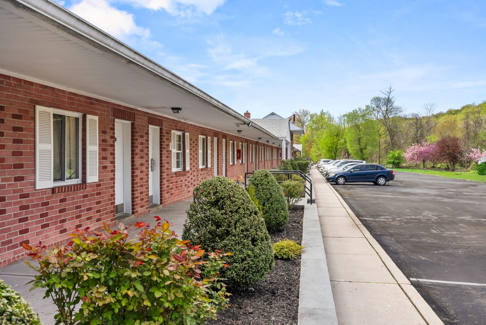 A row of brick motel rooms with white doors, small shrubs lining a concrete walkway, and a parking lot with a blue car in the distance.
