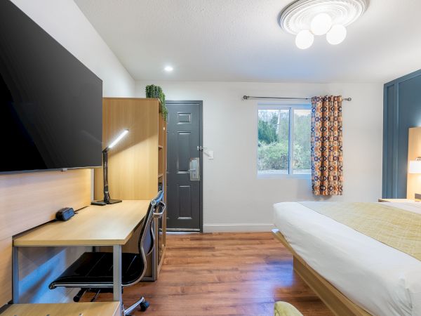 A neat hotel-style bedroom with a desk and chair, a wall-mounted TV, a dark entry door, a window with patterned curtains, and a glowing ceiling light.