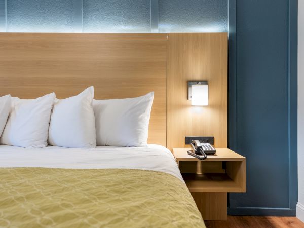 A hotel room with a bed, white pillows, a wooden headboard, a nightstand with a phone, and blue walls with a small wall light.