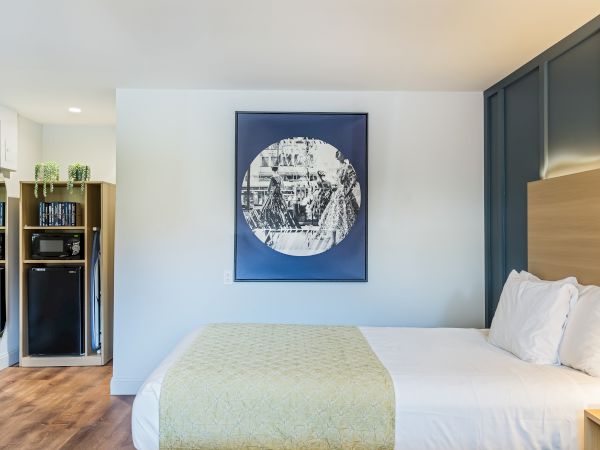 A modern hotel room with a large bed, blue-accent wall art, a wooden headboard, and a small kitchenette area to the left, all in warm lighting.