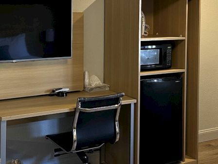 A small home office setup with a desk, swivel chair, wall-mounted TV, and a narrow cabinet containing a microwave and a mini fridge, all in a wood-toned room.