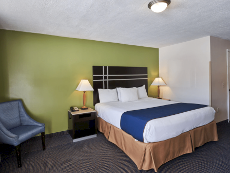 A hotel room with a king bed, green accent wall, nightstands, lamps, a blue bed runner, a beige bed skirt, an armchair, and a doorway in the background.