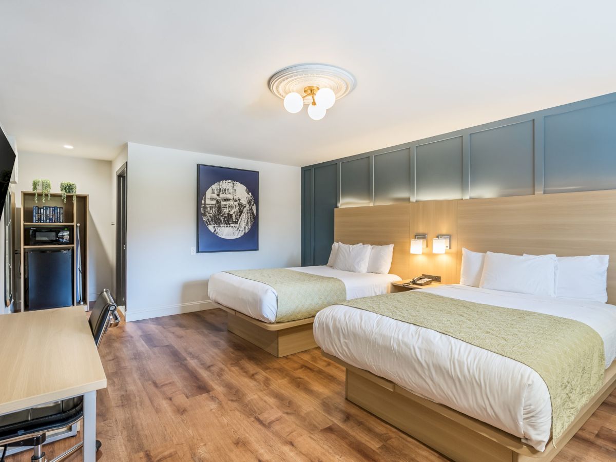 A bright hotel room with two queen beds, a blue padded headboard wall, a wall-mounted TV, desk, and a small kitchenette in the corner, all on warm wood flooring.