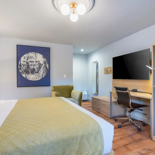 A modern hotel room with a bed, yellow-green bedding, a seating area, desk, wall-mounted TV, and a wooden cabinet; bright lighting and a blue art piece.