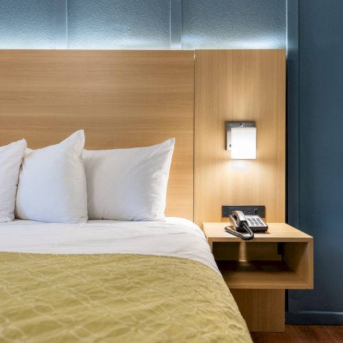 A tidy hotel room with a large bed, white pillows, a mustard bedspread, a wooden headboard, a bedside table, and a wall-mounted lamp by a blue wall.