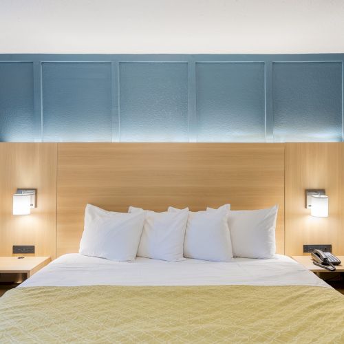 A cozy hotel bedroom with a wooden bed, soft beige bedding, blue paneled walls, and warm ambient lighting, inviting a restful stay.