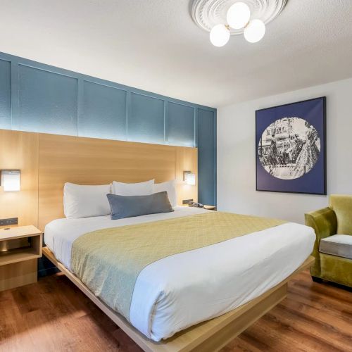A modern hotel room with a king bed, blue upholstered headboard wall, wooden bedside tables, a yellow accent throw, and a green chair, bright lighting.