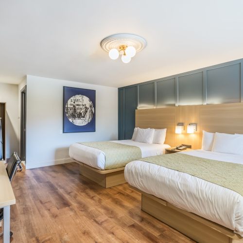 Two plush queen beds with beige throws in a modern hotel room, blue paneled headboard wall, wood floors, desk, wall art, and a TV facing the beds.