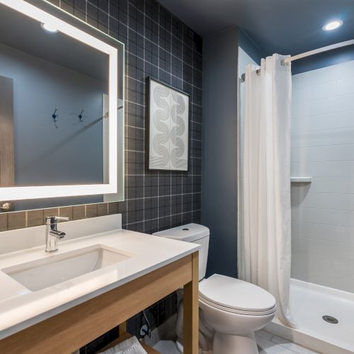 A modern bathroom with a rectangular backlit mirror above a double-sink vanity, dark tiled wall, and a glass-enclosed shower. Ending with a period.