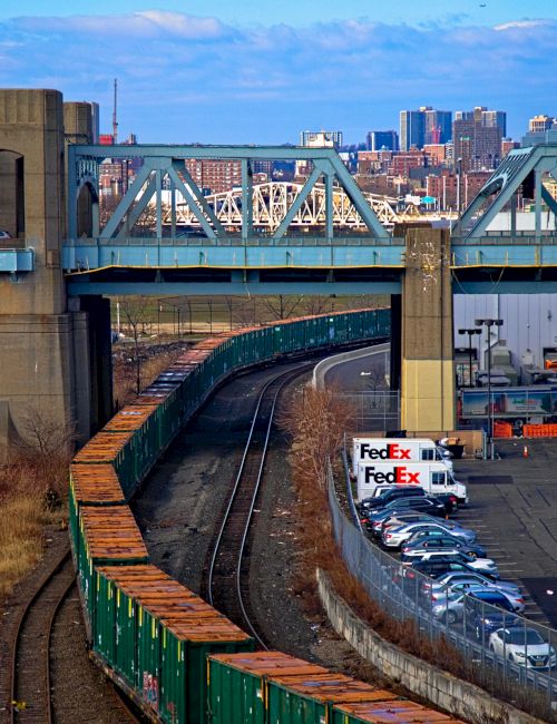 A freight yard beside a curved railway under a blue sky, with a metal bridge overhead and a row of FedEx trucks parked near a building.