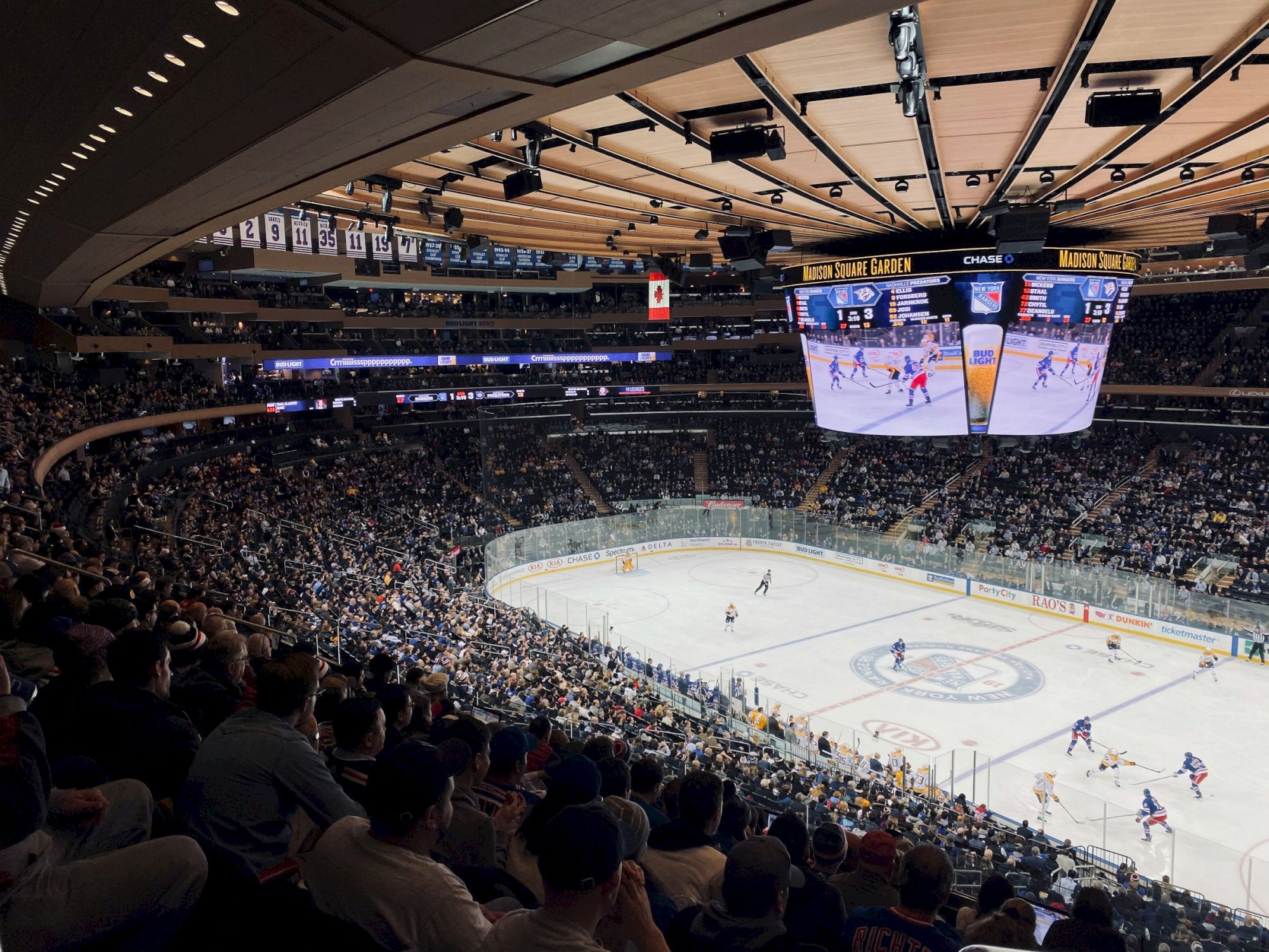 A packed hockey arena with players on the ice, cheering fans, and a large scoreboard hanging above.