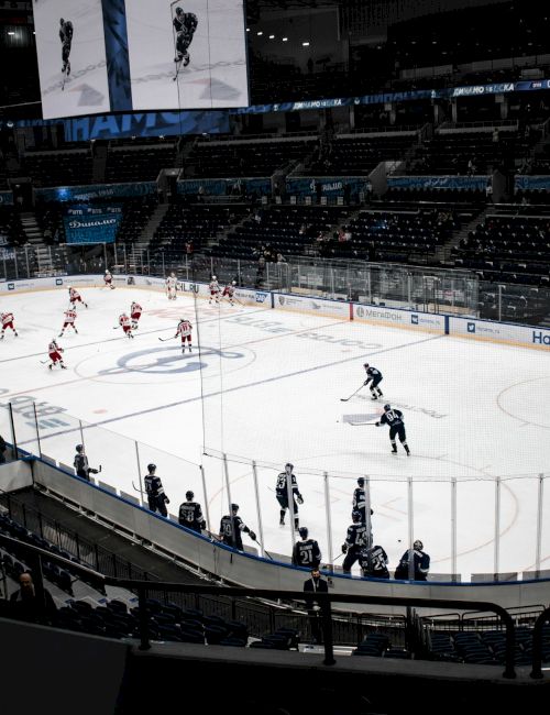 An indoor ice hockey game is underway with players on the rink and spectators in stands, under arena lights, with goal nets and officials on the ice.