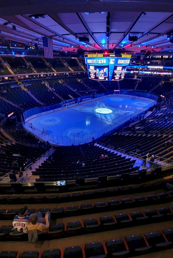 An empty indoor arena with a glowing blue ice rink at the center, surrounded by dark seats and dim lighting, ready for a game tonight.